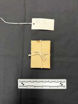 Small light brown cardboard box tied with natural coloured cotton string. This photograph is taken upon a black background, with a scale card and object number included.