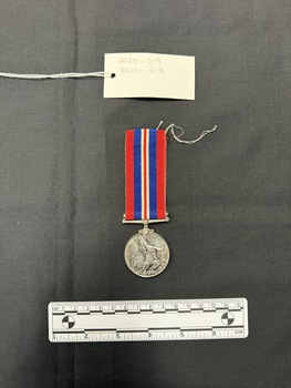 Front view of the War Medal 1939-1945. The silver coloured medal has a central and textured design of the crowned effigy of King George VI, facing left, around the perimeter is the legend 'GEORGIVS VI D:G:BR:OMN:REX ET INDIAE IMP:' The ribbon is coloured in vertical strips: red, blue, white, red, white, blue, red, it is tactile, feeling ribbed under finger. This photograph is taken upon a black background, with a scale card and object number included.