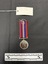 Back view of the War Medal 1939-1945. The silver coloured medal has a lion standing on a double-headed dragon. The ribbon is coloured in vertical strips: red, blue, white, red, white, blue, red, it is tactile, feeling ribbed under finger. This photograph is taken upon a black background, with a scale card and object number included.