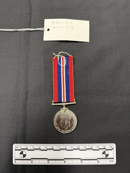 Back view of the War Medal 1939-1945. The silver coloured medal has a lion standing on a double-headed dragon. The ribbon is coloured in vertical strips: red, blue, white, red, white, blue, red, it is tactile, feeling ribbed under finger. This photograph is taken upon a black background, with a scale card and object number included.