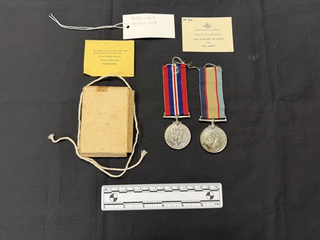 Small cardboard box with natural coloured string alongside two silver medals with coloured ribbed ribbons. Also shown are two pieces of paper, one yellow with black type on it, the other piece a buff coloured heavier stock (card) with dark blue type and a handwritten note in the top left corner in blue ink. This photograph is taken upon a black background, with a scale card and object number included.