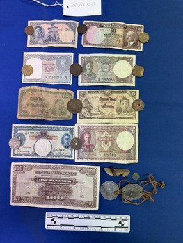 Image shows nine currency paper notes and eleven coins of various sizes and colours from various countries.  There is a set of identity disks, one circular the other octagonal with holes in each and joined by brown leather cord threaded through the holes, the disks also have the name, service number and other details stamped on them, the metal is silver and shiny.  There is also a small wooden shoe shaped like a small heeled shoe or clog and a very small ancient looking coin.