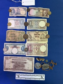 Image shows nine currency paper notes and eleven coins of various sizes and colours from various countries.  There is a set of identity disks, one circular the other octagonal with holes in each and joined by brown leather cord threaded through the holes, the disks also have the name, service number and other details stamped on them, the metal is silver and shiny.  There is also a small wooden shoe shaped like a small heeled shoe or clog and a very small ancient looking coin.