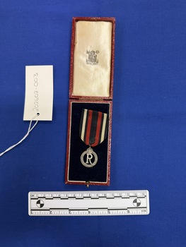 Small silver medal hanging on a piece of red ribbon with a dark blue central stripe and dark blue, white and dark blue side stripes. The medal has a large voided 'R' in the centre with a crown on top, surrounded by the words Queen Alexandra's Imperial Military Nursing Service Reserve. The medal and ribbon sit inside the lower half of a Royal mint box on a blue cushion with the Royal Mint logo stamped on the upper inside and a red covering on the outside of the box.