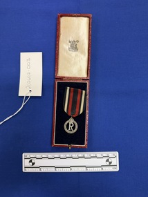 Small silver medal hanging on a piece of red ribbon with a dark blue central stripe and dark blue, white and dark blue side stripes. The medal has a large voided 'R' in the centre with a crown on top, surrounded by the words Queen Alexandra's Imperial Military Nursing Service Reserve. The medal and ribbon sit inside the lower half of a Royal mint box on a blue cushion with the Royal Mint logo stamped on the upper inside and a red covering on the outside of the box.
