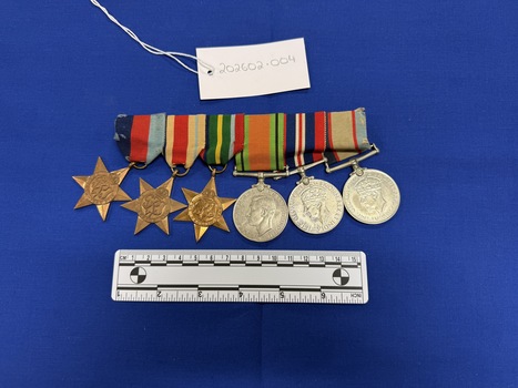 Array of six World War Two medals mounted together on a long pin. 1939-45 Star, Africa Star and Pacific Star are all made from yellow copper zinc alloy, with somewhat sharp 6 points. They each have a central textured design. The Defence Medal, War Medal 1939-45 and Australia Service Medal made from nickel-silver, the fronts have various central and textured designs. Their ribbons are tactile, in various colours including dark blue, red, pale blue, white, green, yellow, pale green, royal blue and black. 