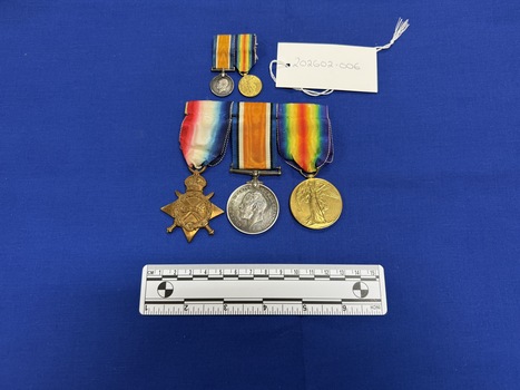 Three World War One medals mounted on a badge pin together. The 1914-1915 Star a bronze four pointed star with crown at top with a watered ribbon of red merging into white which merges into blue. The British War Medal 1914-1918 shows a bareheaded effigy of King George V facing left, ribbon is orange with borders in dark blue, black and white. The Victory Medal, 1914-1919 is bronze with a winged figure of Victory on the obverse, the ribbon has a 2 rainbow design, with the violet from each rainbow on the outside edges moving through to a central red stripe where both rainbows meet.  There is also a miniature replica pair of the British War Medal and the Victory Medal.