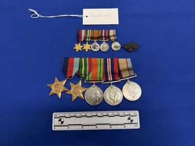 Array of five World War Two medals mounted together on a long pin. 1939-45 Star and Pacific Star are all made from yellow copper zinc alloy, with somewhat sharp 6 points. They each have a central textured design. The Defence Medal, War Medal 1939-45 and Australia Service Medal made from nickel-silver, the fronts have various central and textured designs. Their ribbons are tactile, in various colours including dark blue, red, pale blue, white, green, yellow, orange, pale green, royal blue and black.  There is also a set of matching miniatures and a WWII metal badge with two loops on the reverse and the front embossed with army, navy and airforce emblems topped with a crown and set on a boomerang, and 'RETURNED FROM ACTIVE SERVICE' in text.