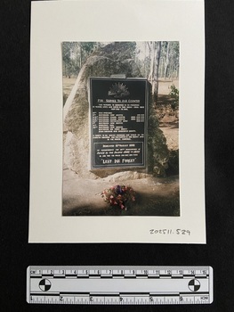 Coloured photograph of a large memorial plaque mounted on a tall rock, trees behind in the distance. A floral wreath has been placed at its base.