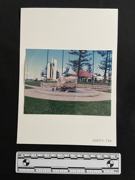 Coloured photograph of a stone memorial set in a pond with a concrete edge and surrounded by small stones. The pond is on flat grassy land and behind it are stone steps for seating. In the background are trees and a pavillion.