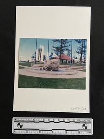 Coloured photograph of a stone memorial set in a pond with a concrete edge and surrounded by small stones. The pond is on flat grassy land and behind it are stone steps for seating. In the background are trees and a pavillion.