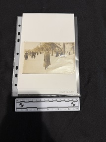 Photographic postcard depicting people walking along a tree lined avenue.  There is a woman walking towards and smiling at the camera wearing a long coat, dress and hat, holding something in her left hand.