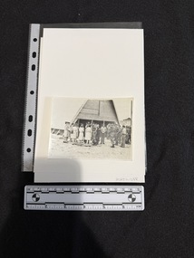 Photograph depicts a large group of people standing in front of a triangular shape building, a chapel.  Most of the people on the left of the photograph are women in uniforms comprised of dresses just below knee length with dark shoes and hats of various designs.  Most of the men depicted are in uniforms of long pants, shirts with sleeves rolled up and most are wearing hats.  There is an elderly man in the middle of the crowd in a black religious outfit that has a white collar and a long sash dangling near his knees.
