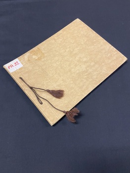 Fawn manilla folder type cardboard cover with a line of ornate embossed pattern running vertically down the left side (two inces in from the spine). Brown thread is tied through the two hole punches on the left side with both ends finished with a knot and tassle.
There is a white sticker label in the top left corner with 'PA32' in red marker.