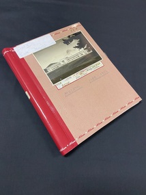 Light pink photograph album with feint, thin red stripes running vertically down the cover. The red spine continues an inch inwards on the left edge. The word 'Album' is repeated continuously around the top and bottom of the cover (in red text across the pink background and white text across the red spine). There is a white label stuck to the spine and to the top front cover. A black and white postcard image of a building has been taped on the front with the text 'Malaya 1955-1956' handwritten below in blue pen ink.