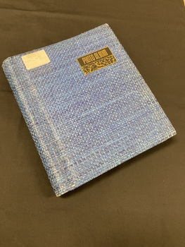 Blue woven covered photograph album with a black plate attached to the upper right cover that reads photo album in gold lettering and an onate pattern below the words (plate measures 1x3inch). A white label is attached to the top left corner with WWI and II inscripted on it.