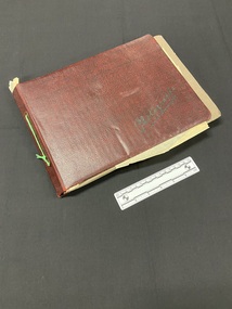Red and black mottled cover with the word photographs scrawled across the bottom left corner in black print. The album is tied together with green string through the spine. The brown cardboard pages inside the cover protrude on the right side and the bottom. The album is quite worn.