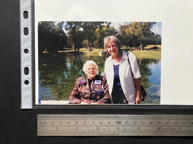 A 6x4 colour glossy photograph of two women, one elderly and seated with white hair wearing medals on the left side of her chest, the other older woman with grey hair standing wearing a white unbuttoned shirt over a navy blue top.  The background is a small lake and parklands.