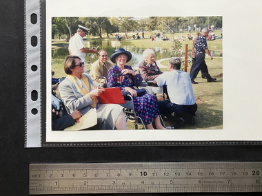 Glossy finish, colour photograph of group of people sitting and standing by a pond in a park. There are two elderly women seated near the centre, a man is squatting in front of one of them, back to the photographer, there are others seated and standing around and in the background.