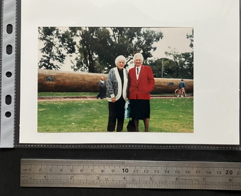 Glossy finish, 4x6 colour photograph of two women, from left to right, Wilma Young (neé Oram) and Vivian Statham (neé Bullwinkel) standing in a park. In the background a man on a bike is reading one of the plaques along a long horizontal tree trunk; another man is looking at the women being photographed.