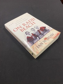White paperback book. At the top of the cover are three small black planes flying to the left of the sub-heading. Below this is the title printed in large red letters and underneath is a picture of three smiling Australian nurses standing close together, the two nurses at either end have red capes and the rest of the photo is in black and white.
Directly below the image is a line of black barbed wire and at the bottom of the page is a feint image of soldiers drawing fire towards a ship and the red Japanese sun symbol overlayed across the top of the image.