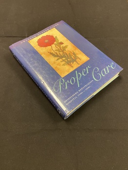 Hardback book with a dark blue dustjacket. Along the spine the title is printed in light aqua script, followed by the printed text of the sub-title and author's name, there is a mark on the bottom of the spine where a sticker has previously been removed.
The front shows an image of a red flower with some greenery set in a light brown rectangle, below which the title is again printed in light aqua script.