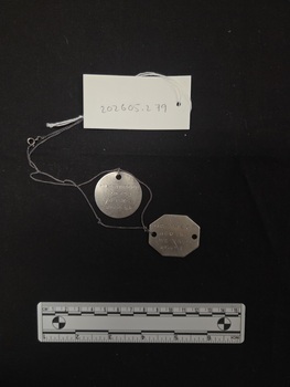 Two thin, aluminium identification discs strung on a fine metal chain. One is octagonal in shape, one is round. Lettering is engraved on the surface of both tags.
