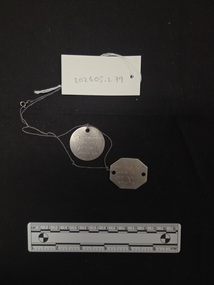 Two thin, aluminium identification discs strung on a fine metal chain. One is octagonal in shape, one is round. Lettering is engraved on the surface of both tags.