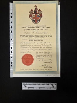 Buff card certificate with a blue border, red seal embossed with the coat of arms of Melbourne, printed black text and two signatures. The coat of arms of Melbourne is printed in colour at the top of the certificate. 