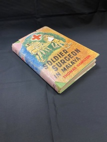 Green hard-cover with dust-jacket.The cover is orange and green with a red medical cross at the top and a drawing of wounded soldiers being assisted by medics, the title is below in black and the author's name in red. The cover image continues around the spine.