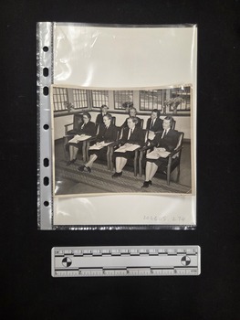 Black and white photograph of seven women in uniform seated in two rows. The four women in the front row have folders of paperwork open in their laps. 