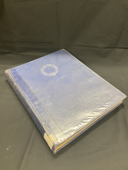 Plastic covered dark blue hard-cover book. Title and author in silver at the top of the spine. White spine label sticker at the base. Front cover has a circular emblem with Latin text printed in silver in the top centre of the cover.