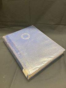 Plastic covered dark blue hard-cover book. Title and author in silver at the top of the spine. White spine label sticker at the base. Front cover has a circular emblem with Latin text printed in silver in the top centre of the cover.