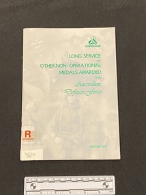Staple bound booklet with opaque cover containing green metallic writing over card cover collage of photographs of military persons and equipment. There is a reference sticker on the front and a spine label sticker below it.