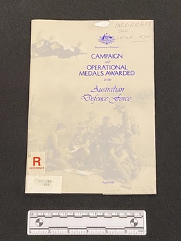Staple bound booklet with opaque cover containing blue metallic writing over card cover collage of photographs of military persons and equipment. There is a reference sticker on the front and a spine label sticker below it.