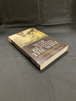The bottom half of the cover and the spine are black with the title printed in silver. The top half is a sepia photograph of two smiling prisoners of war, the bottom of the image is overlaid with black barbed wire.