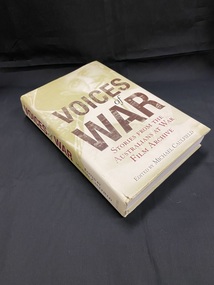 Hard-cover book with dust-jacket. The image on the dustjacket is pale green and wraps around both front and back covers and spine, it shows the face of a soldier and of men walking across a wooden pathway across mud and water. The title is printed across the middle of the front cover in lettering made up of black and white photos of a variety of faces.