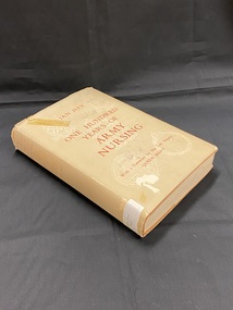 Tan hardcover book with a lighter dust jacket. Front cover has title and author printed in red, There are five white images of badges from different Nursing Services positioned across the front and back covers.