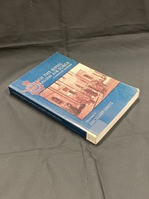 Dark green paperback with spine label taped to base of spine. Title printed in white down the spine. Front cover has title and series information printed in white, overlaying an Australian Air Force emblem. In the centre of the front cover is a sepia photograph image of 13 men in long coats standing out the front of a large stone building.