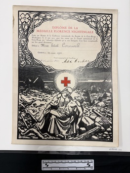 A large board mounted copy of a certificate that has black print artwork of a woman holding a man on her lap in a landscape that has broken wood, ground and a half sunken ship present.  The middle of the certificate has a red cross in a white circle and the top half has text bordered with thorns and foliage.