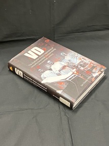 Black dust-jacket over black hard-cover book. Spine shows the gold Australian Army emblem at the top and then the title in white, there is a spine label at the base. Front cover has VD in large white letters at the top, followed by the sub-title and an image of three men in surgical gowns in a surgical room. The page has translucent red bubble images over it.