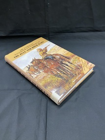 Brown hard-cover book with a light brown dust-jacket displaying a large illustration of a soldier aiming a rifle, sitting on horseback beside another horse in open fields. The title is printed in black across the top and down the spine. There is a spine label at the base of the spine.