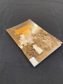 Small paperback with an orange spine with title printed down it and a white spine label taped to the bottom. The front cover shows a sepia toned image of a group of wounded soldiers on crutches and in wheelchairs with a nurse standing amongst them. There are two orange templates in the top centre of the page with the title information printed inside them in black print.