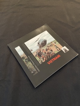 Square paperback book with a black cover. Red printed title on spine and cover. Front cover has a long thin photo image of jungle foliage down the left side, next to which is part of the title printed vertically down the page. On the right side is a photo of soldiers in a field with two helicopters taking off beside them and the word Vietnam printed in red text beneath.