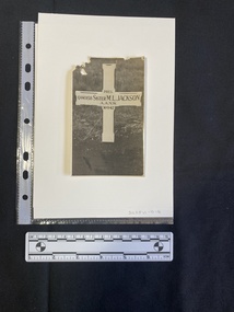 A black and white photo of a headstone in the shape of a cross with an inscription.