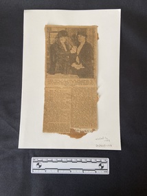 Newspaper clipping with roughly cut edges. Black and white photograph at the top of a woman pinning a medal on a second woman. Below are two columns of typed text.