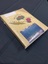 Cream coloured photo album with illustrations of a gum nut, gum leaf, and a waratah flower and matching words. At the bottom is a photo of a memorial wall with wreaths and bouquets of flowers.