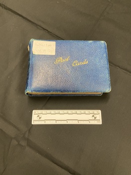 Small blue photograph album with worn cover. White sticker label in top left corner with 'WW1 VAD' written on it. 'Post Cards' is printed in gold in the centre of the front cover.