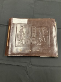 Brown leather cover bound with a tied leather strip down the left side. Embossed Egyptian image on the front cover. White sticker label in top left corner.