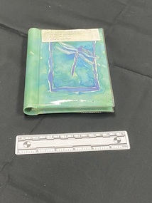 Small, green, plastic covered photo album with a blue watercolour dragonfly inside a blue border on the front cover. Large white sticker label across the top.